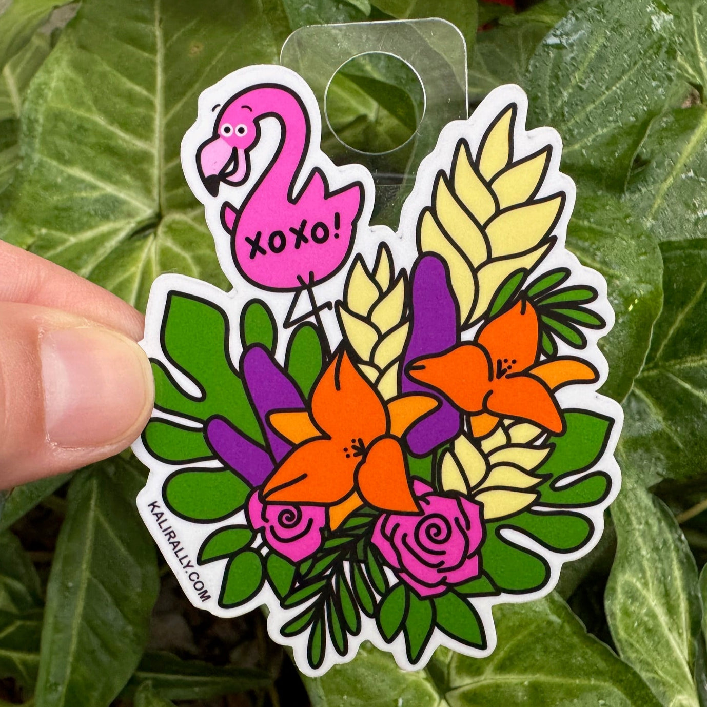 Tropical Bouquet Sticker with a flamingo and floral design held over green leaves, by Kalirally