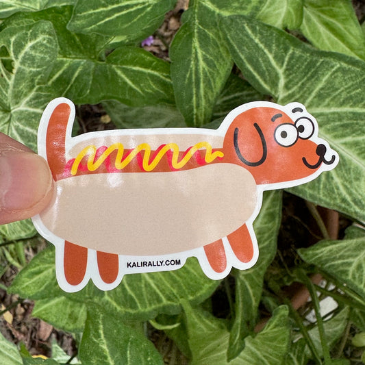 Glossy vinyl Doxie Hot Dog Sticker featuring cartoon dachshund as a hot dog with ketchup and mustard, displayed in front of a plant, by Kalirally