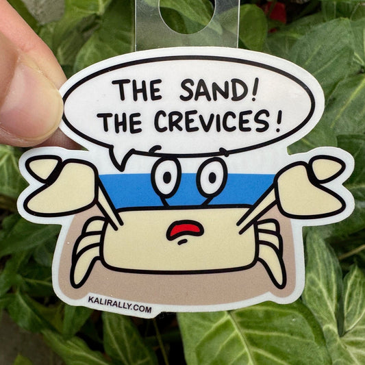A picture of our cartoon Sand Crab sticker with text 'The sand! The crevices!' held over green leaves.