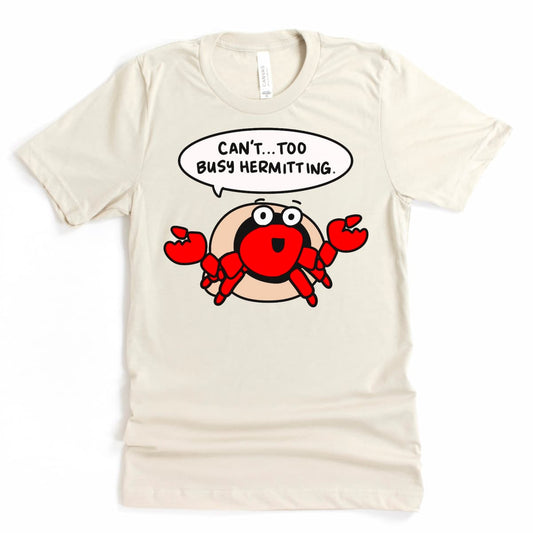 Flat lay of a Heather Dust Colored Beach Tee Shirt - “Can't. Too Busy Hermitting.” t-shirt by Kalirally, featuring a cartoon hermit crab design for introverts.