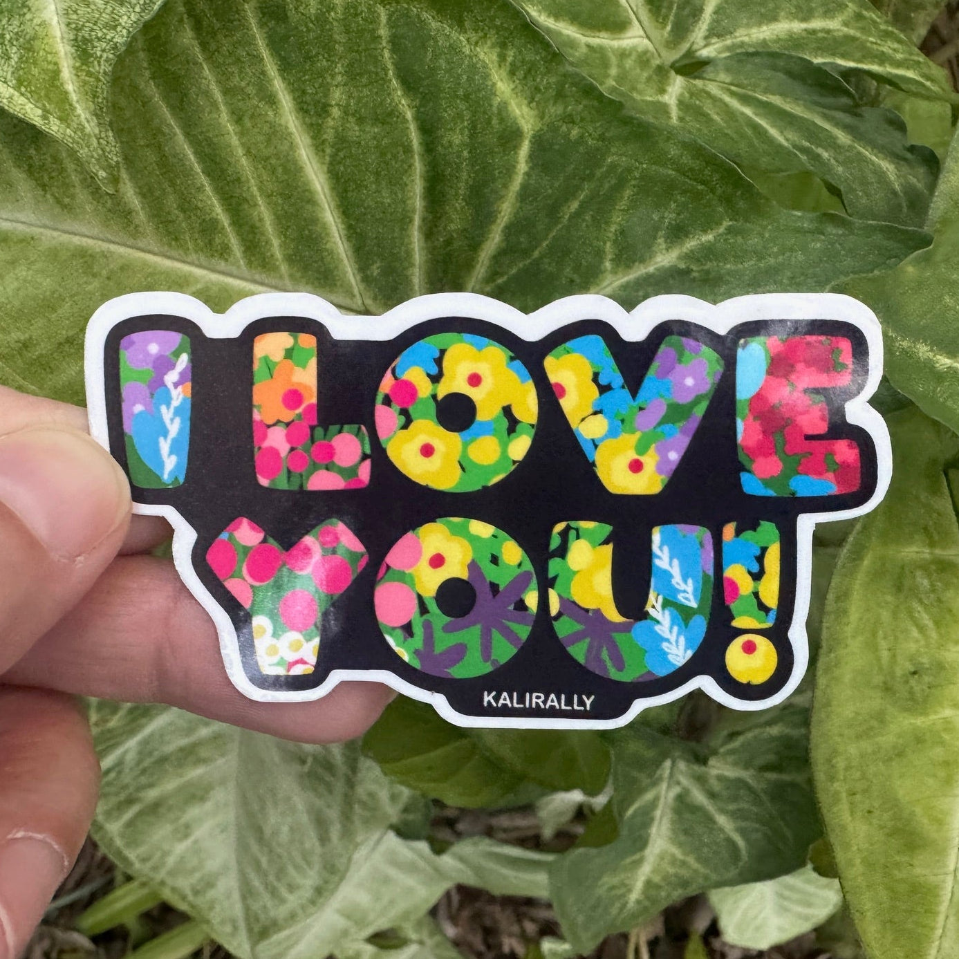 Hand holding a colorful 'I LOVE YOU!' sticker against a leafy green background