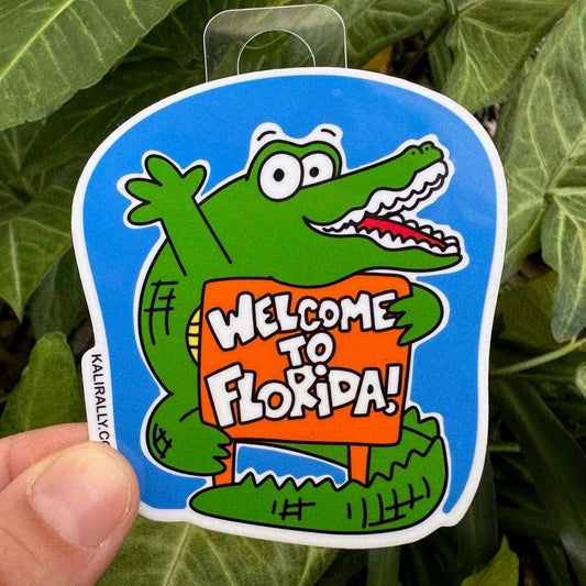 Cartoon alligator hugging a colorful “Welcome to Florida!” sign – fun vinyl Florida sticker souvenir held in front of green leaves, by Kalirally